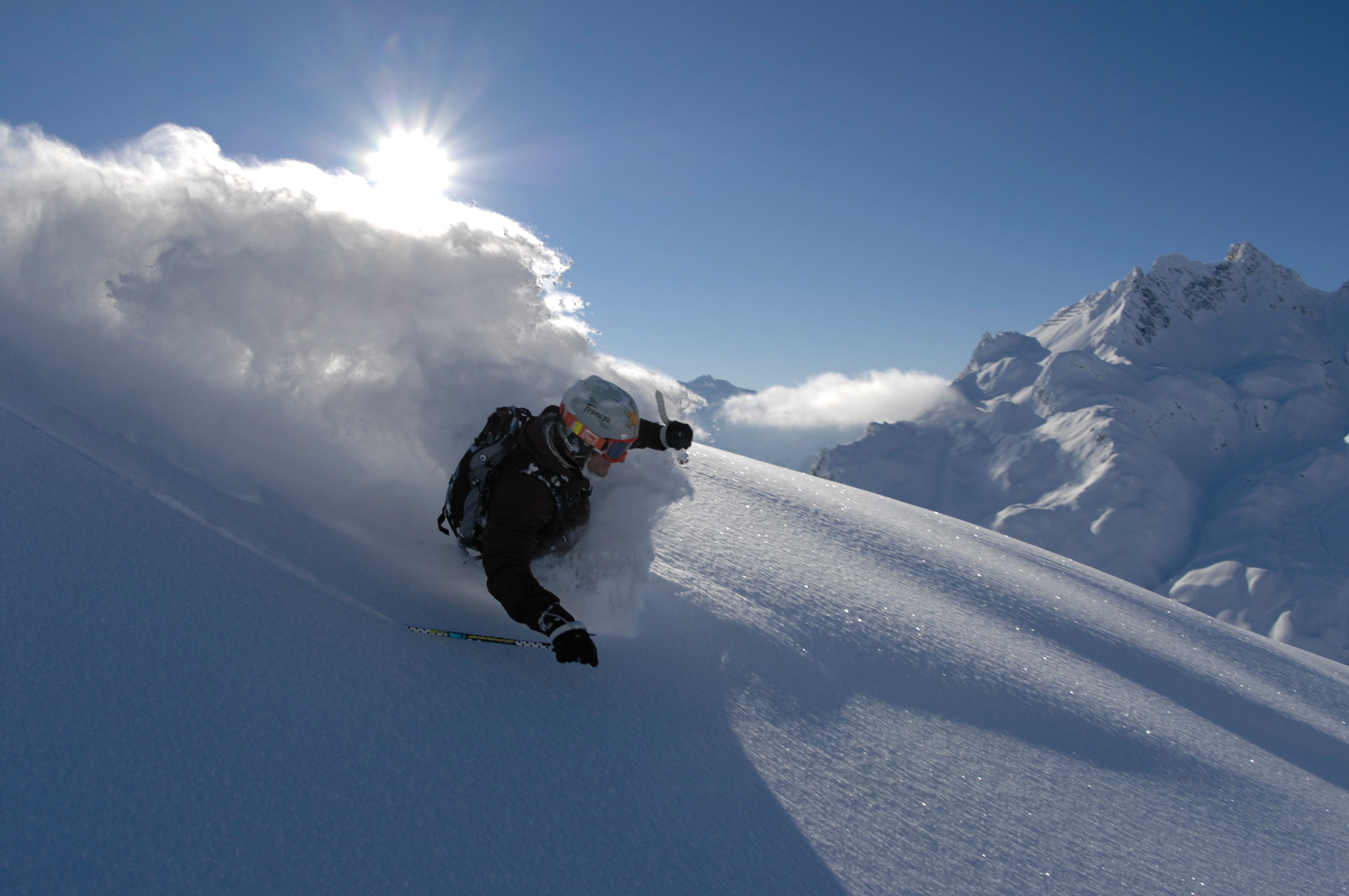 En person står på ski ned ad en sneklædt skråning med en klar himmel og bjerge i baggrunden.