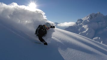 En person står på ski ned ad en sneklædt skråning med en klar himmel og bjerge i baggrunden.
