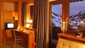 A cozy room with a wooden desk, a chair, a television, a window with a view of snow-covered mountains, and a bedside table with a lamp and books. - Bergheim (Lech am Arlberg)