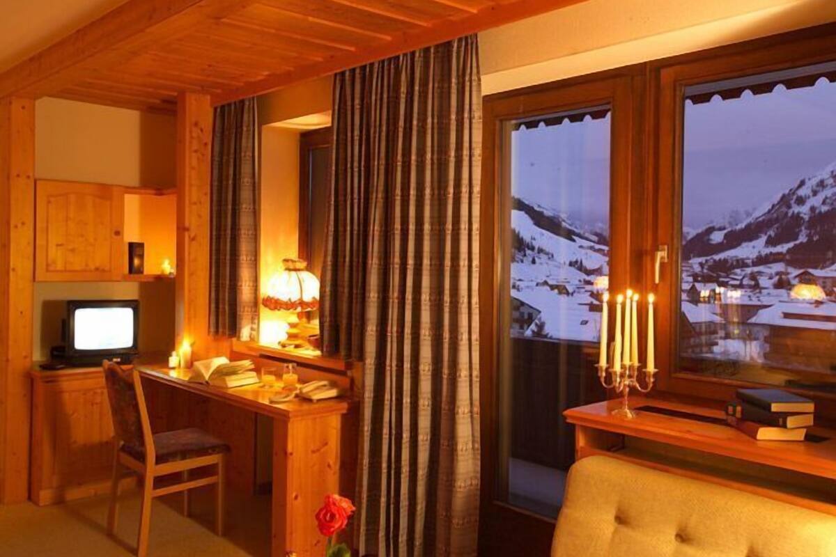 A cozy room with a wooden desk, a chair, a television, a window with a view of snow-covered mountains, and a bedside table with a lamp and books.