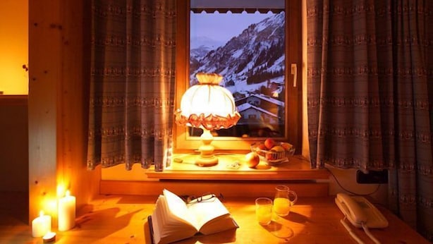 A cozy room with a window overlooking a snowy mountain, a table with a lamp, candles, and a book. - Bergheim (Lech am Arlberg)