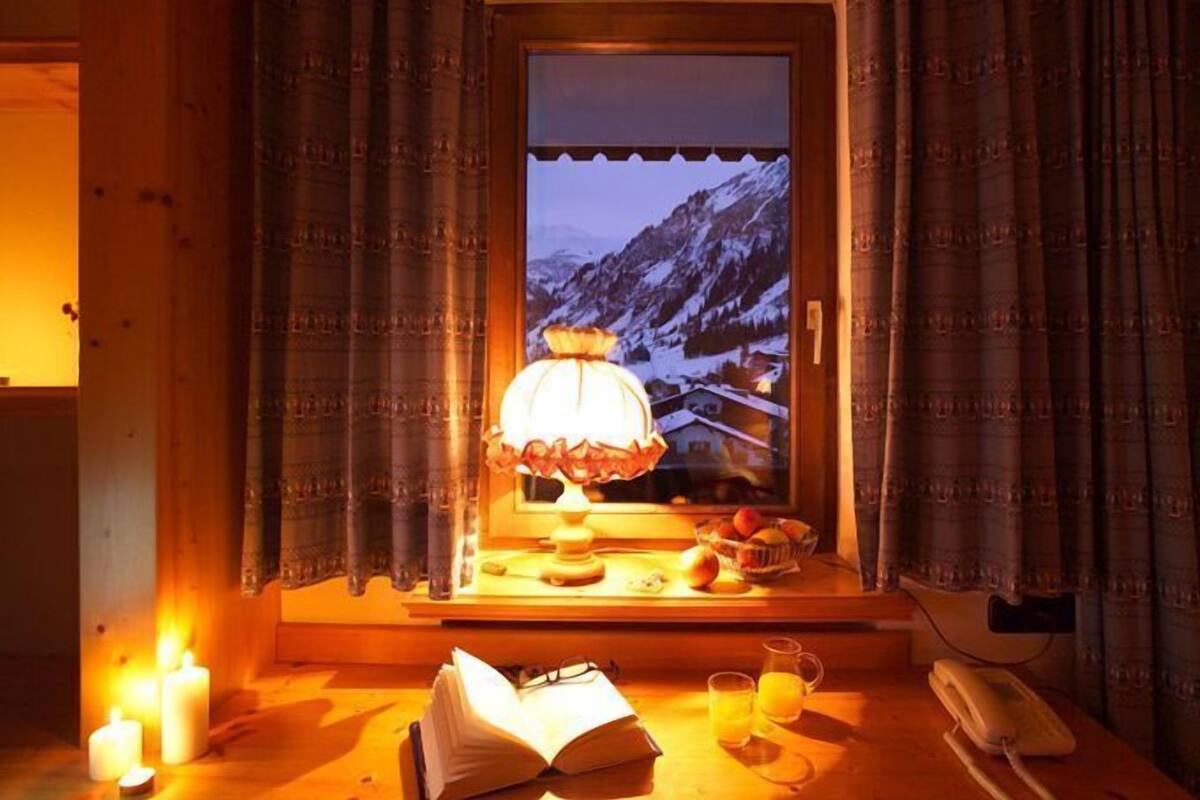 A cozy room with a window overlooking a snowy mountain, a table with a lamp, candles, and a book.