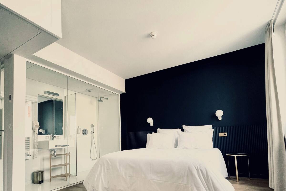 A modern hotel room with a large bed, a walk-in closet, a bathroom with a shower, and a dark accent wall.