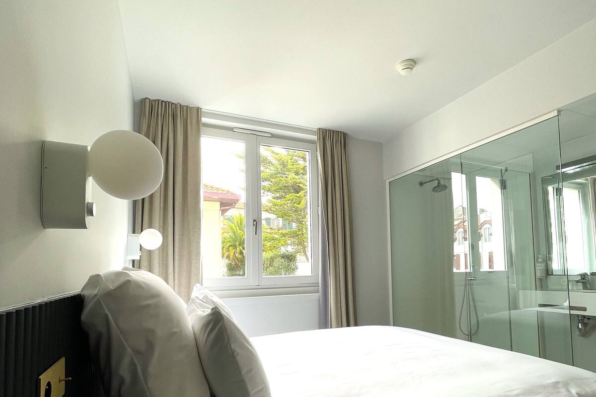 A hotel room with a bed, a window with curtains, a shower area, and a wall-mounted light fixture.