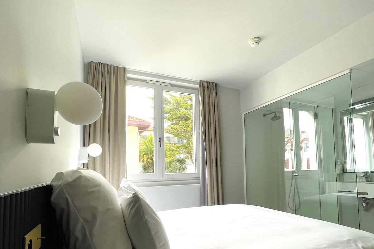 A hotel room with a bed, a window with curtains, a wall-mounted light, and a glass shower enclosure.