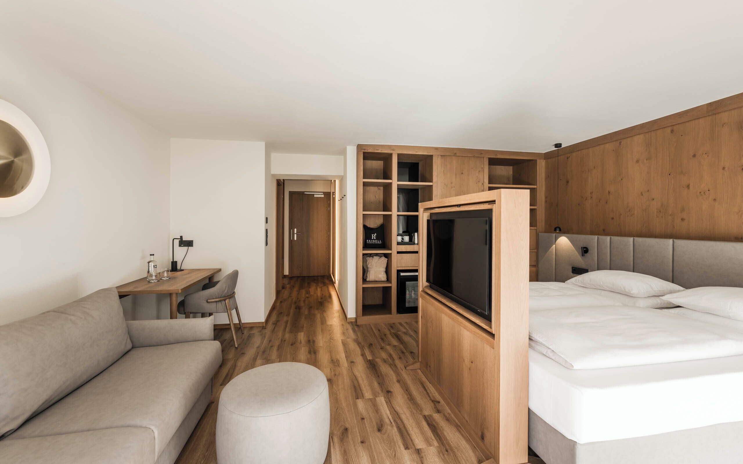 A modern hotel room with a sofa, a TV, a bed, a wooden wardrobe, and a desk.