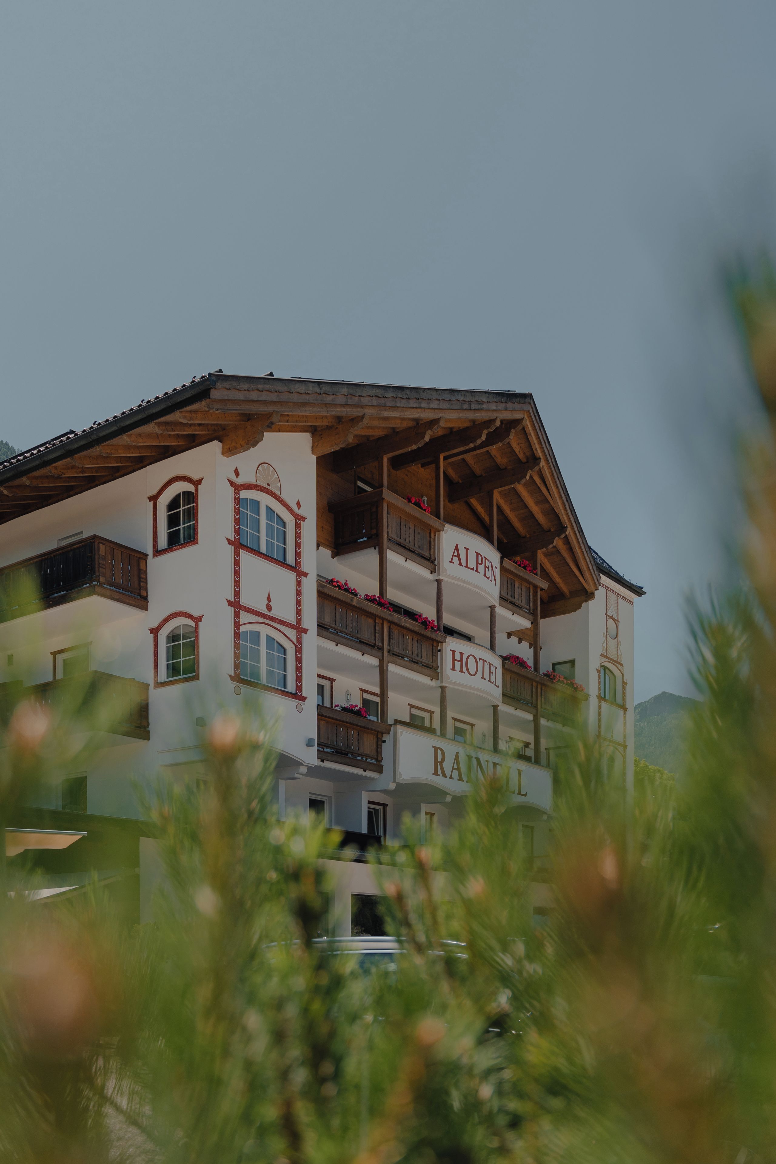 Un hotel de varios pisos con balcones, un cartel que dice 'ALPEN HOTEL' y un paisaje montañoso de fondo.