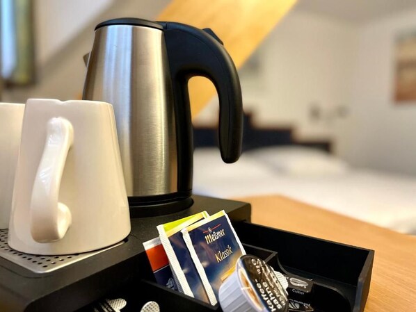 A hotel room with a bed, a bedside table with a kettle, a tray with a coffee pod, a remote control, and a mug. - Insel Hotel (Cologne)
