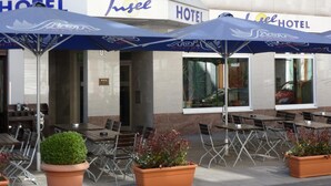 An outdoor seating area with tables and chairs, covered by blue umbrellas, and a building with 'Insee Hotel' signage. - Insel Hotel (Cologne)