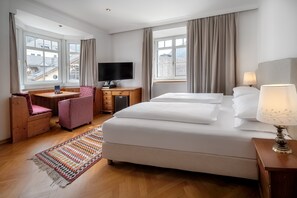 A hotel room with a bed, desk, chair, TV, and windows. - Classic Hotel am Stetteneck (Ortisei)