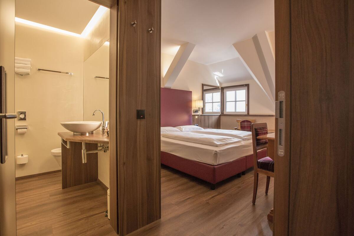 A modern bedroom with a large bed, a bedside table, and a chair. There is a bathroom with a sink and toilet. The room has wooden floors and a sloped ceiling.