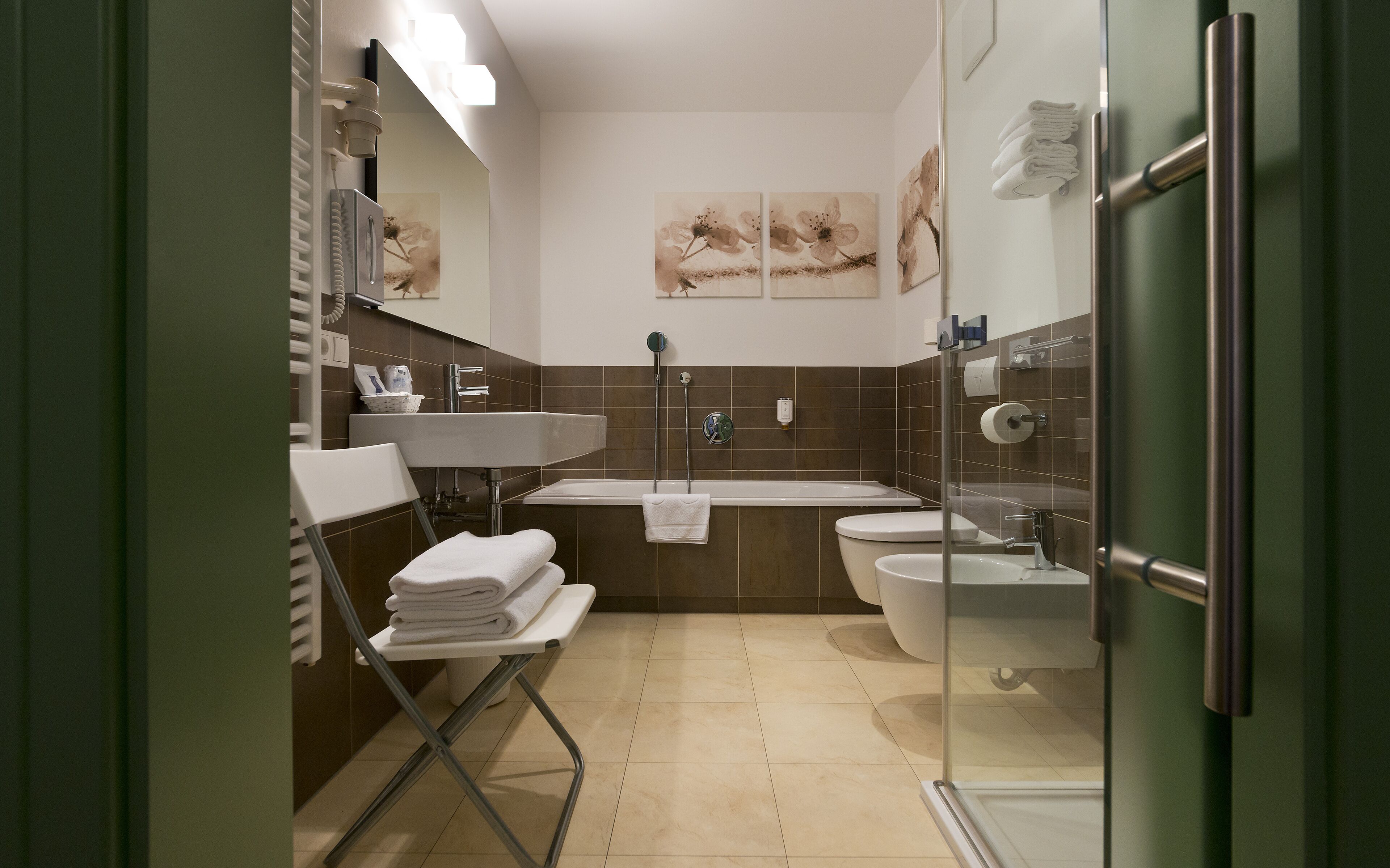 A modern bathroom with a glass shower enclosure, a white sink, and a toilet. There are towels stacked on a chair and a mirror above the sink.