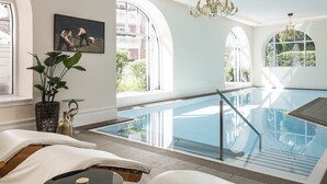 Une piscine intérieure moderne avec des parois en verre transparent, offrant une vue sur l’extérieur, et un espace de détente confortable agrémenté d’une plante et d’un tableau.