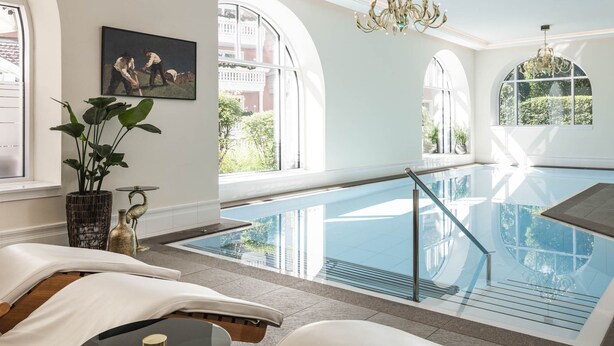 Een modern binnenzwembad met glazen wanden, uitzicht naar buiten en een comfortabele zithoek met een plant en een schilderij.