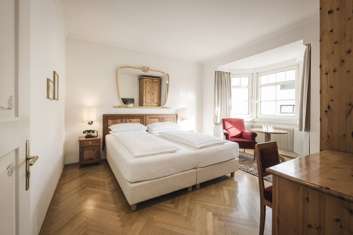A hotel room with a large bed, a wooden desk, a red armchair, and a large window.