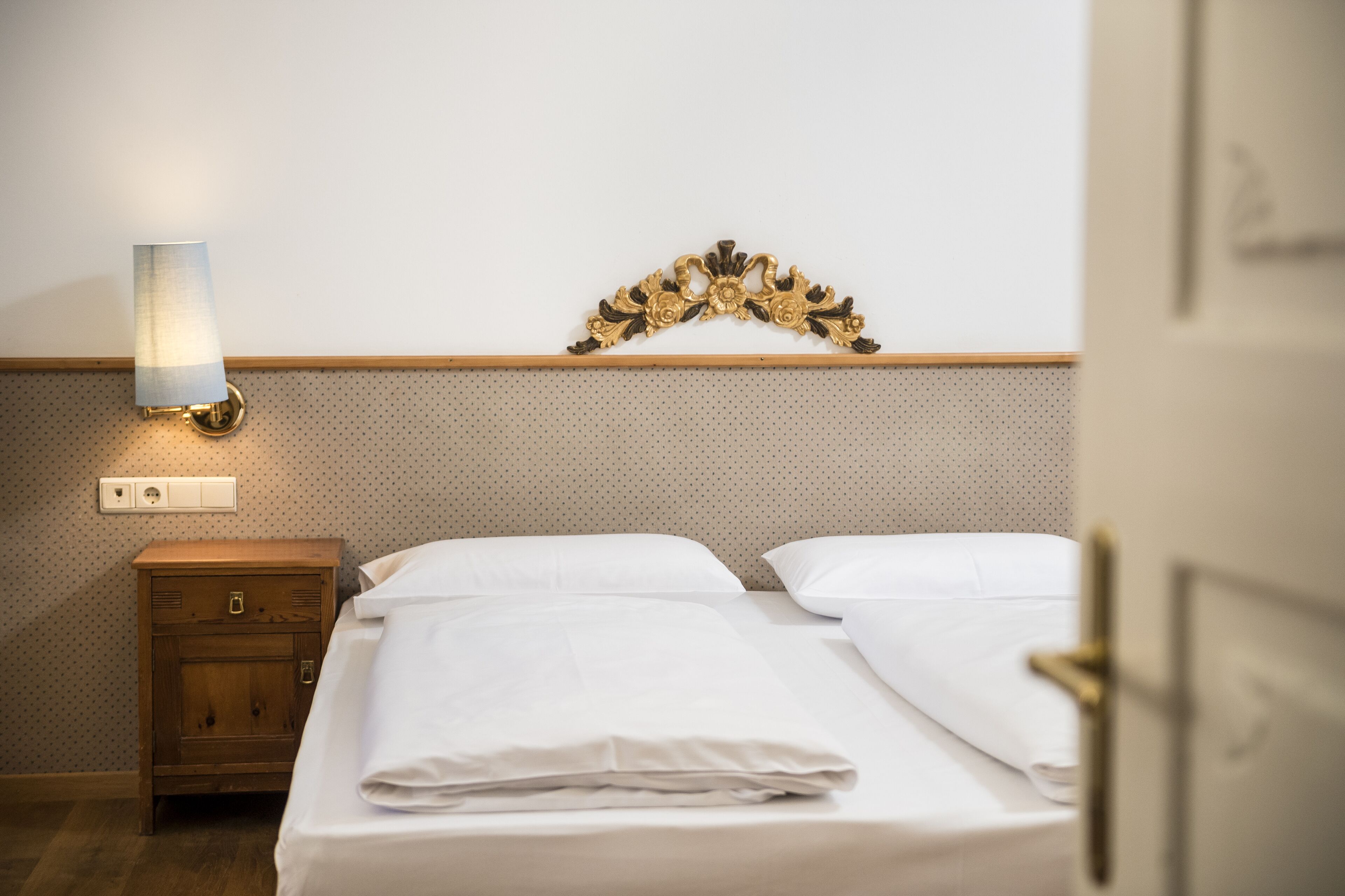 Un lit bien fait, avec du linge de lit blanc, une table de chevet en bois surmontée d’une lampe, et un élément décoratif accroché au mur au-dessus du lit.