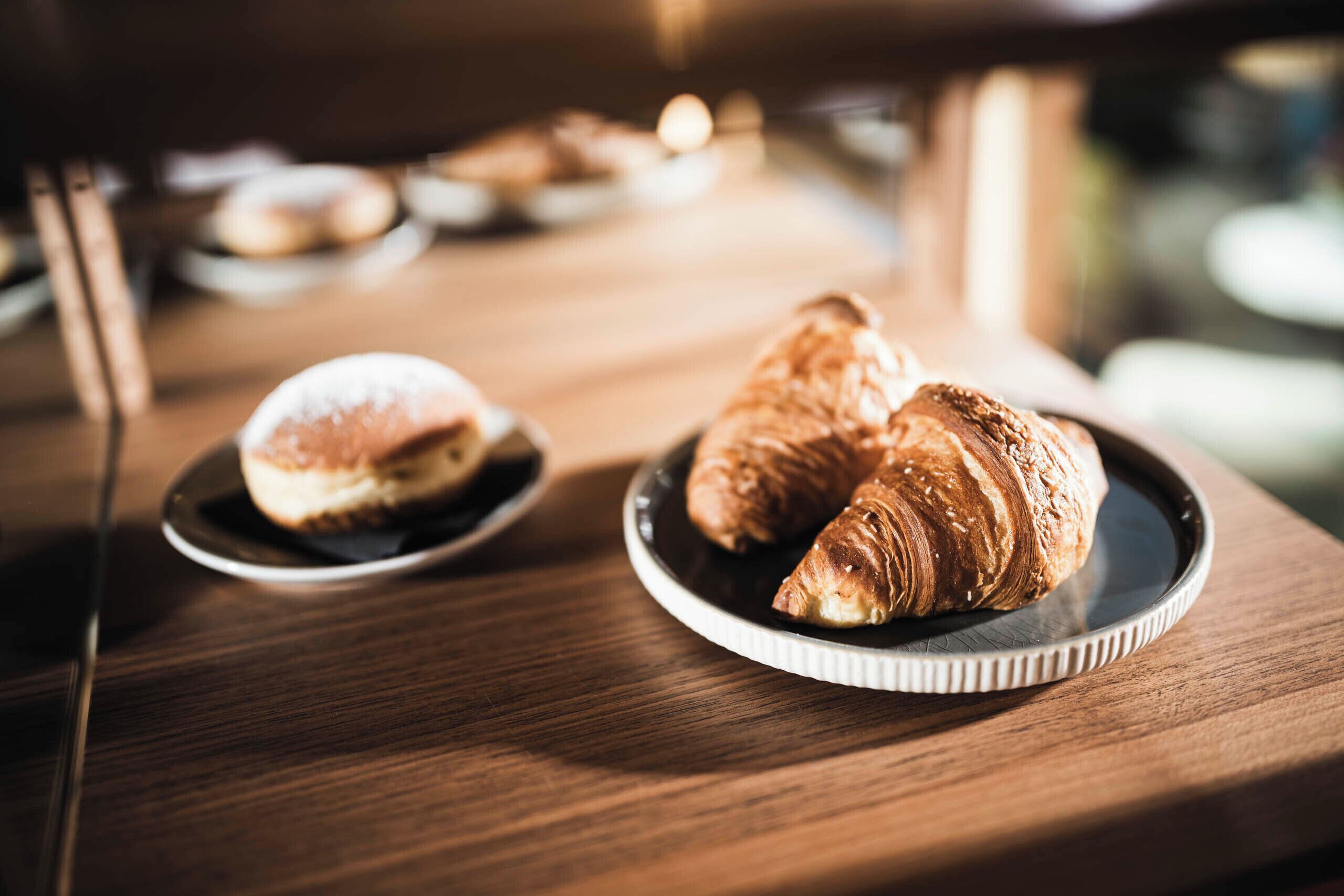 A wooden table with two plates of pastries, one with a round pastry and the other with a croissant.
