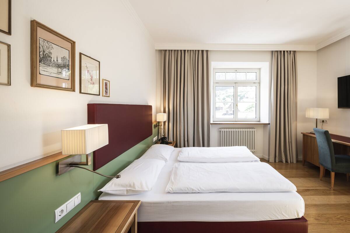 A hotel room with a bed, bedside lamps, a desk, a chair, a television, and framed pictures on the wall.