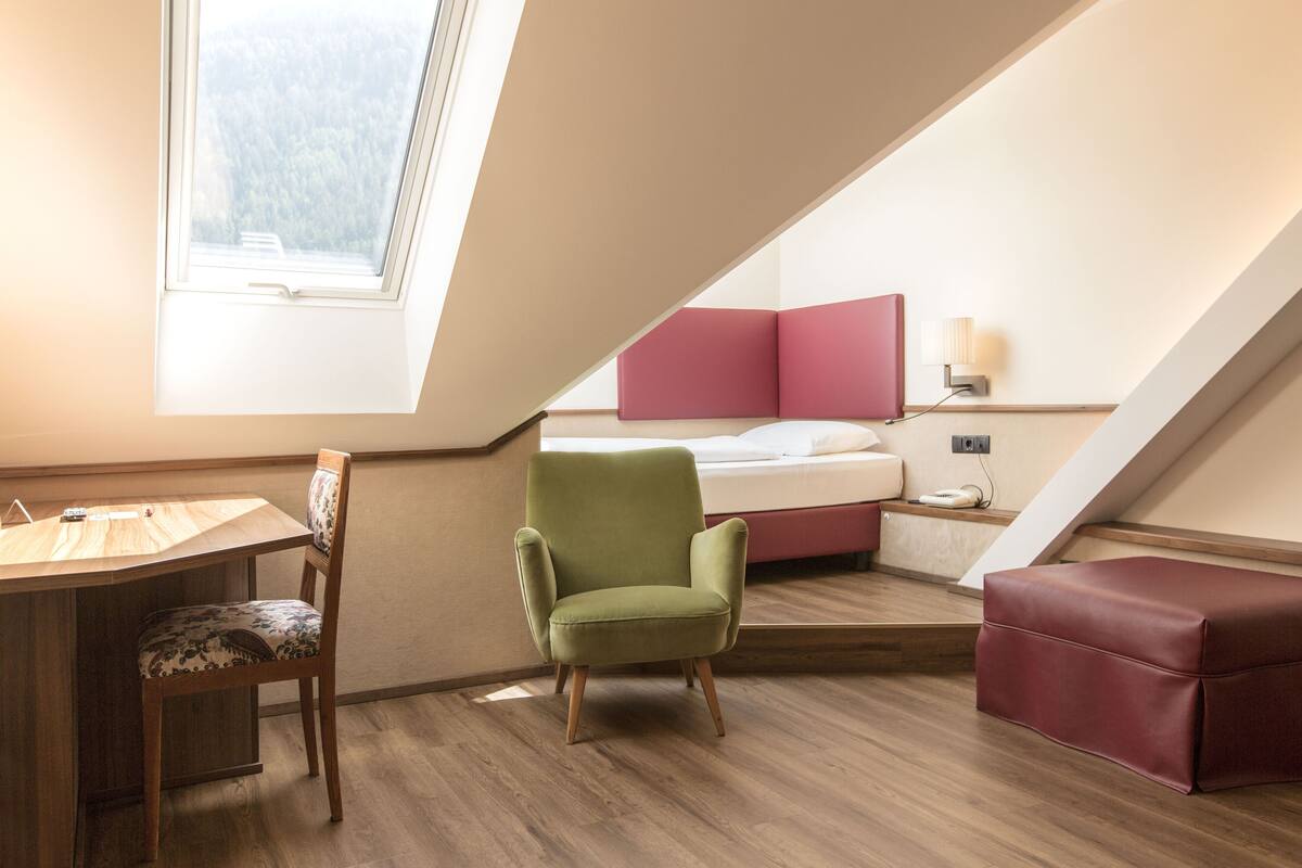 A room with a sloped ceiling, a desk, a chair, a bed with pink headboard, and a red ottoman.
