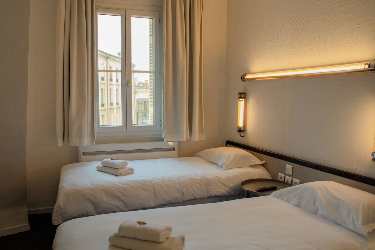 A hotel room with two beds, a window with curtains, and wall-mounted lights.