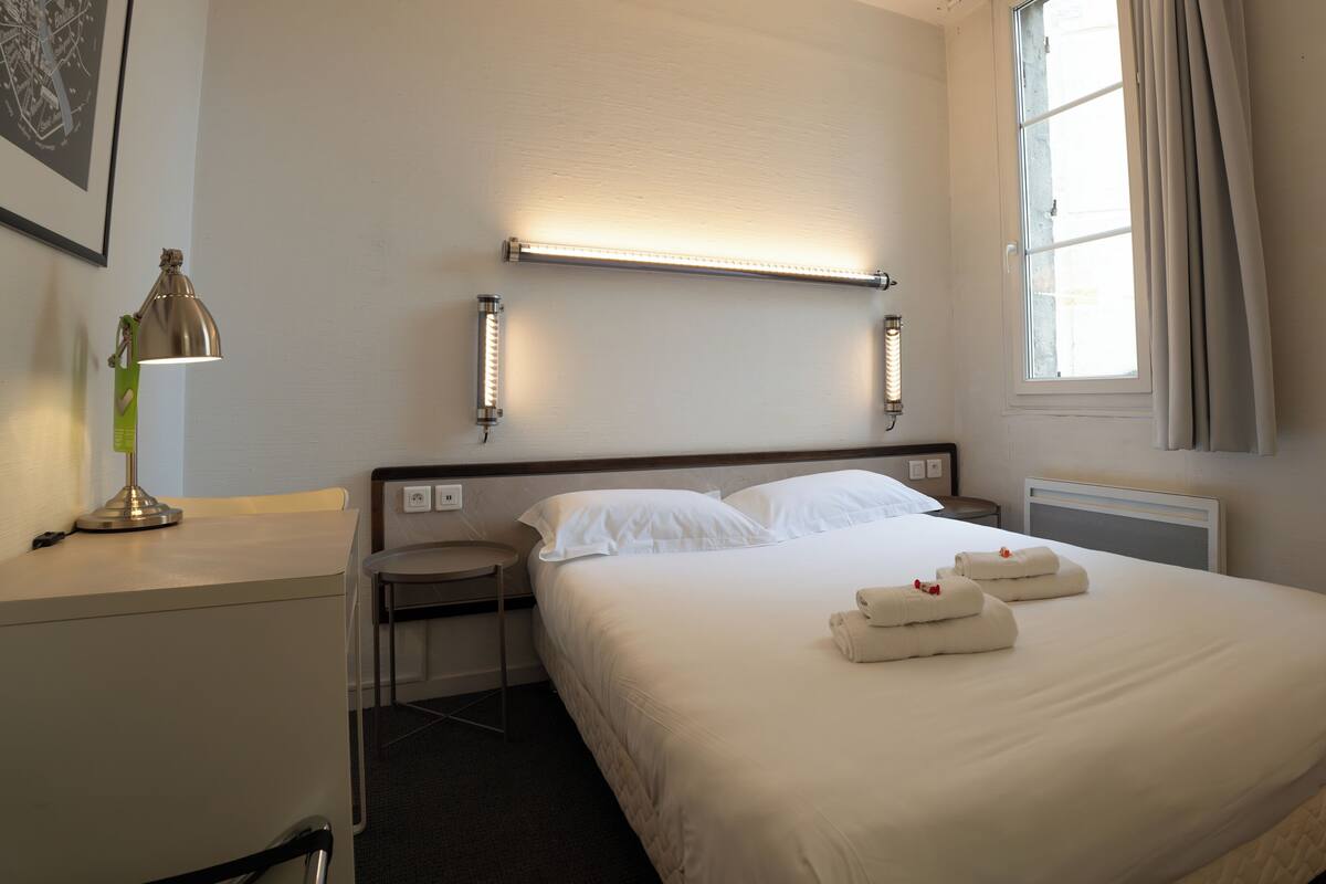 A hotel room with a bed, bedside table, lamp, and a wall-mounted light fixture.