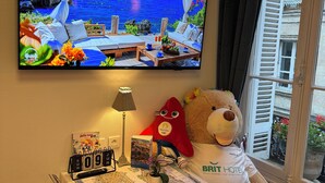 A room with a television displaying a scenic view, a table with a calendar, a lamp, a teddy bear, and a window with a view of a building. - Hotel des Grands Hommes - Bordeaux Centre (Bordeaux)