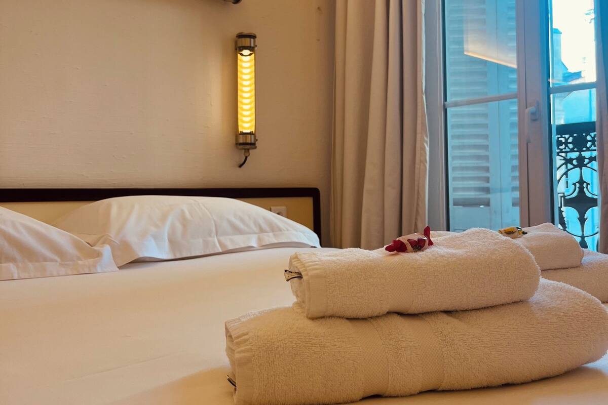A hotel room with a neatly made bed, a wall-mounted light, and a view of the city through a window.