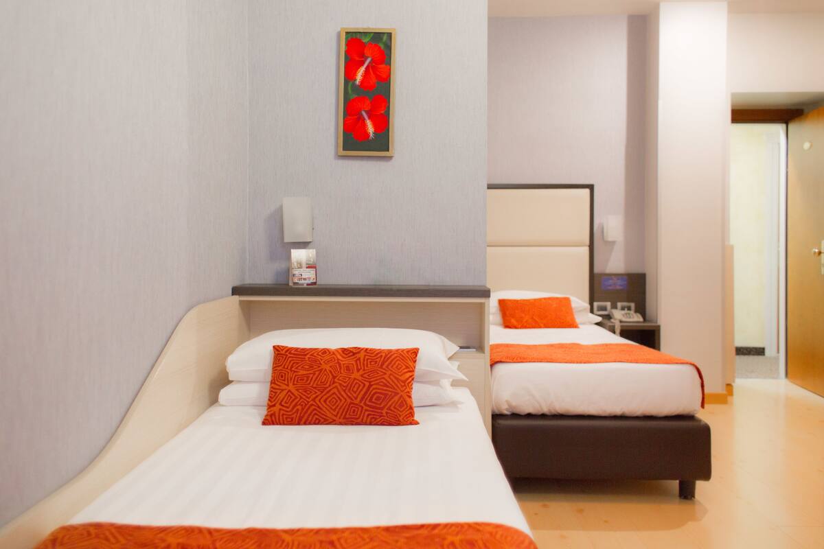 A hotel room with two beds, each with white bedding and orange accents. There is a small table with a telephone and a wall-mounted light.
