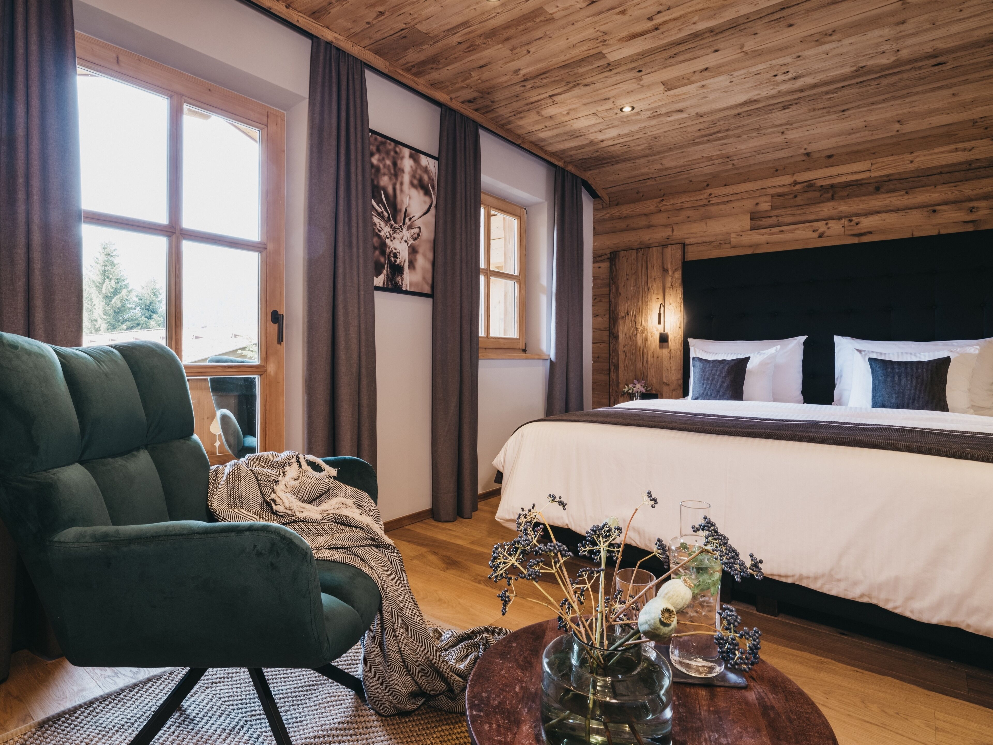 A bedroom with a wooden ceiling, a large bed with white and dark pillows, a green armchair, a wooden table with a vase of dried flowers, and a window with curtains.