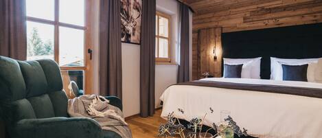 A bedroom with a wooden ceiling, a large bed with white and dark pillows, a green armchair, a wooden table with a vase of dried flowers, and a window with curtains.