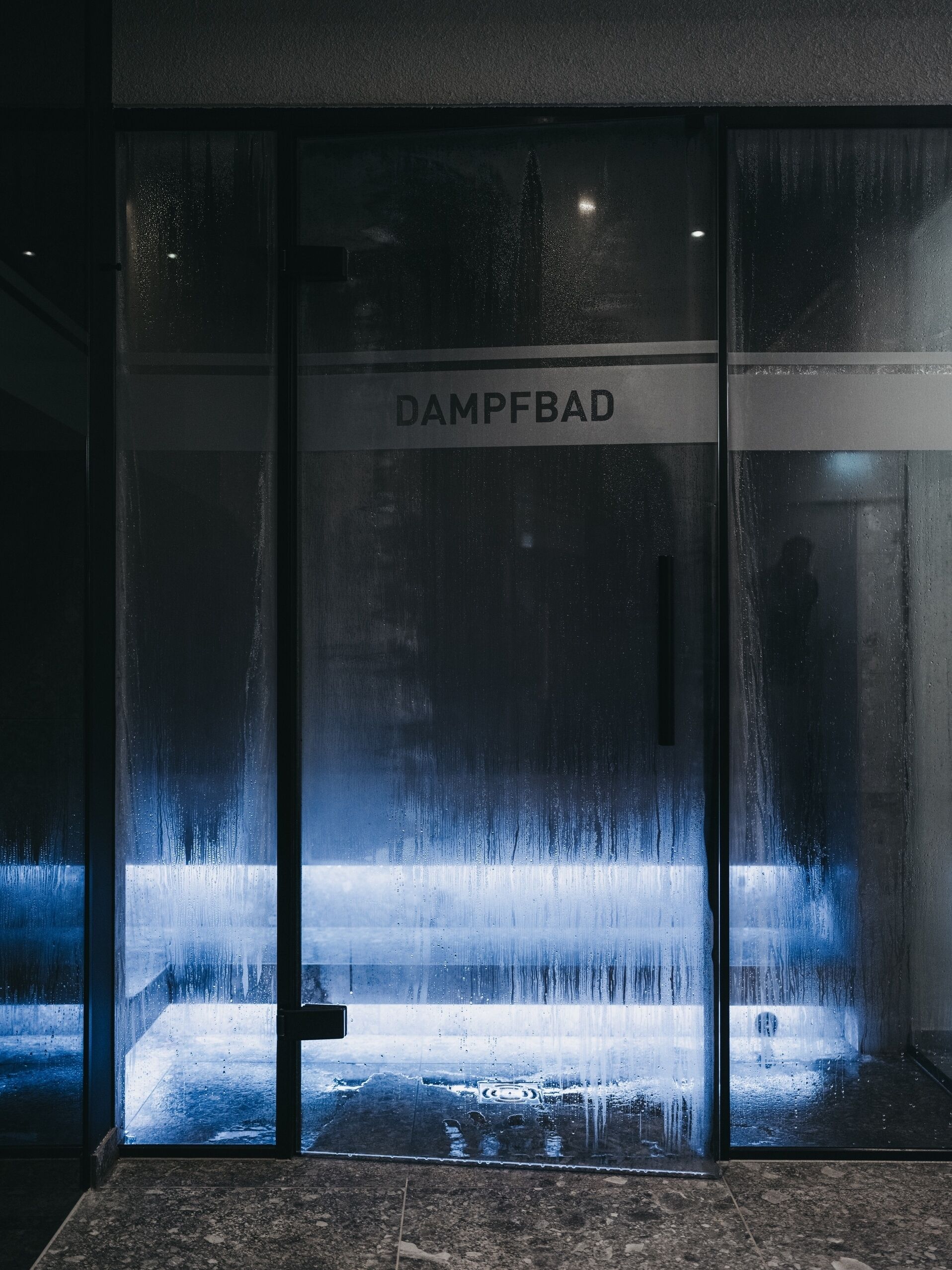 Une porte vitrée portant l’inscription « DAMPFBAD » et donnant sur un trottoir mouillé.