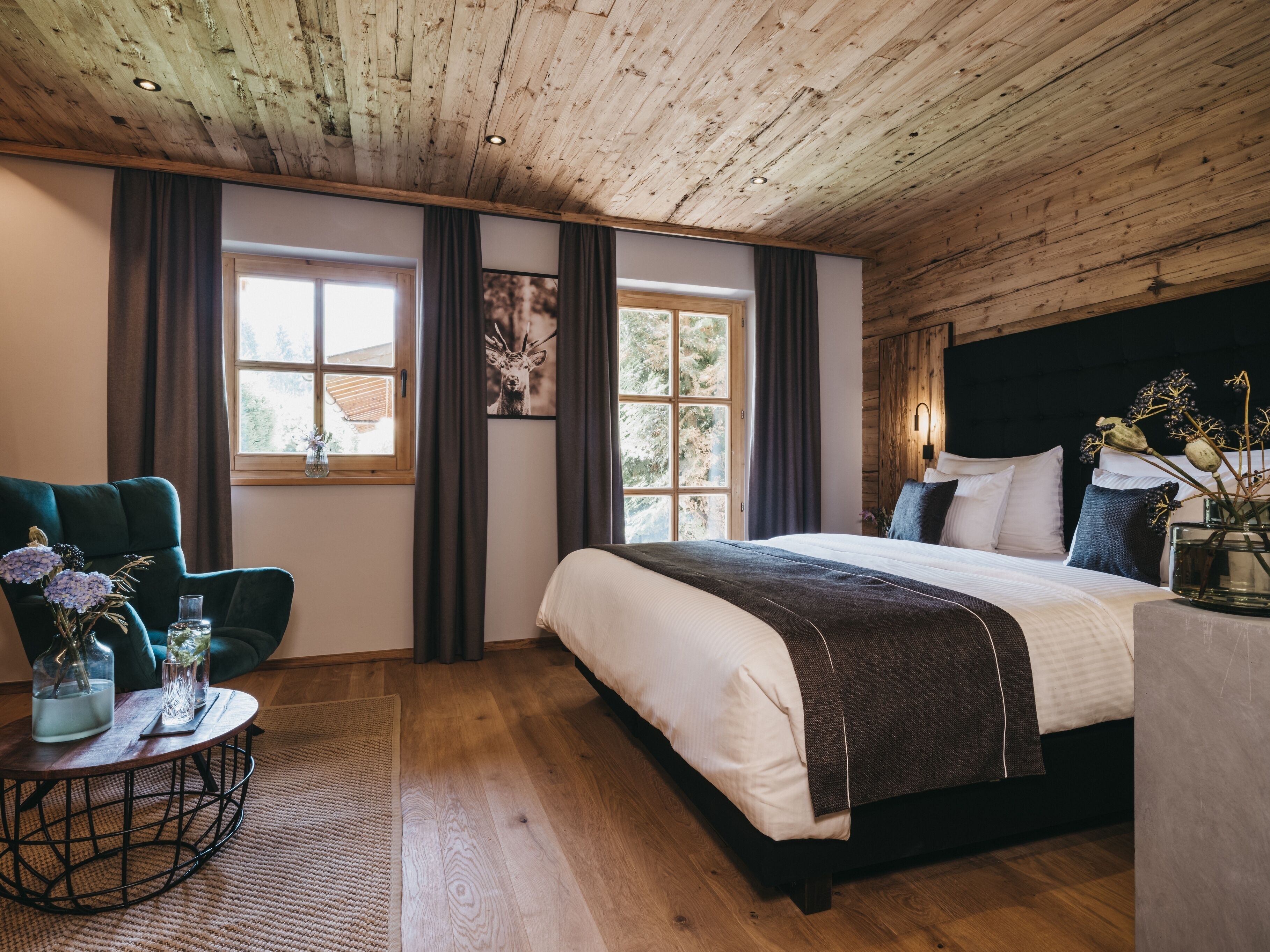 A bedroom with a wooden ceiling, a large bed with white bedding, a green armchair, and a small round table with decorative items.