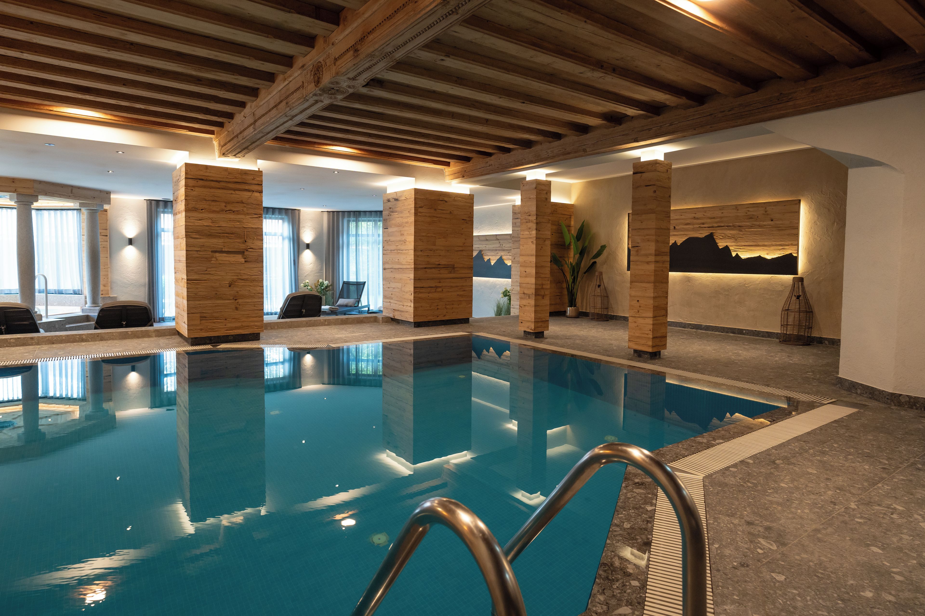 Una piscina interior con pilares de madera, vistas a montañas a través de una ventana y un área de descanso con sillas.