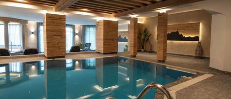 Une piscine intérieure avec des piliers en bois, une vue sur les montagnes par une fenêtre et un espace détente avec des fauteuils.