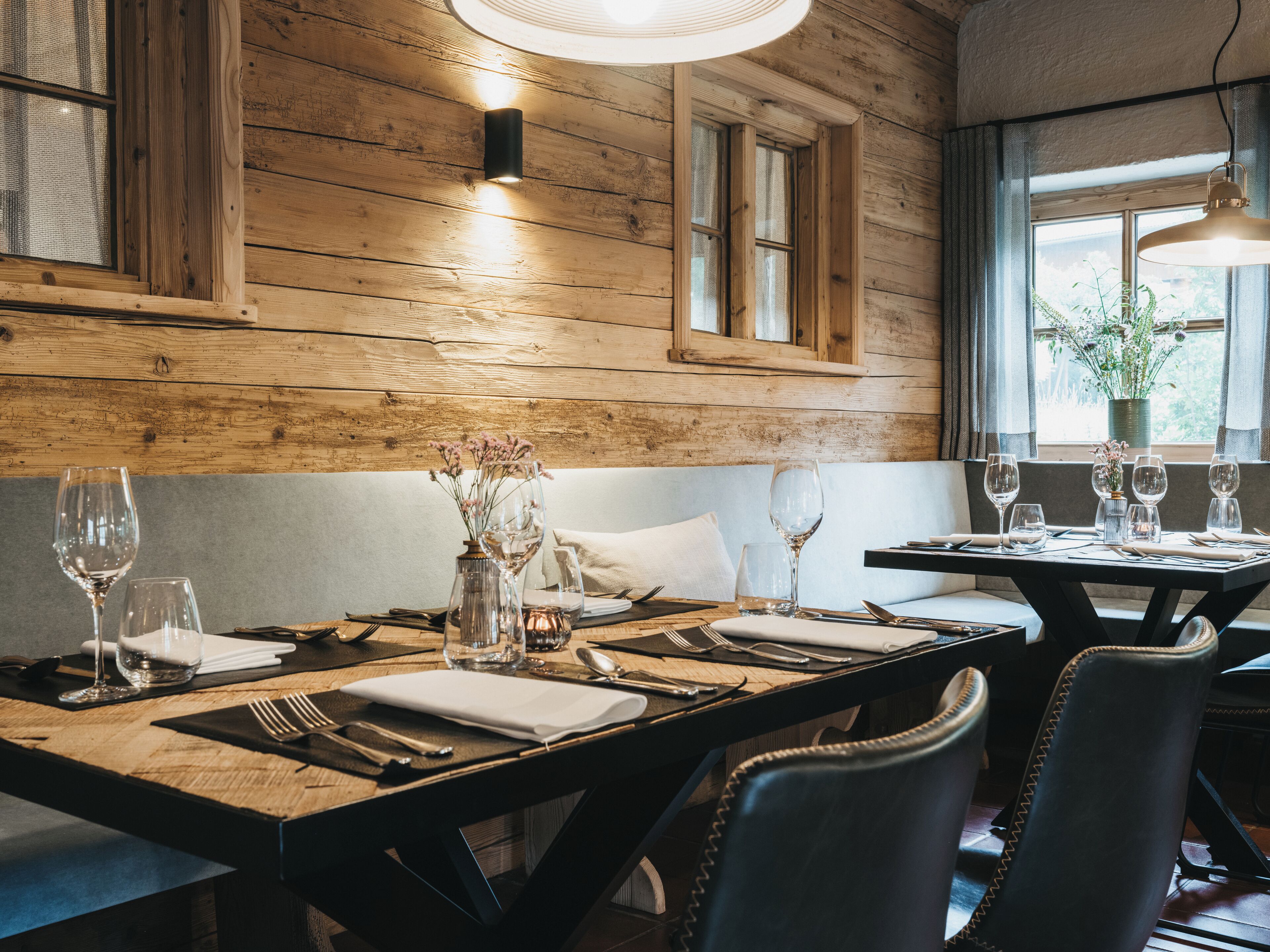 A wooden interior with a dining table set for two, featuring a vase with flowers, clear glasses, and cutlery.