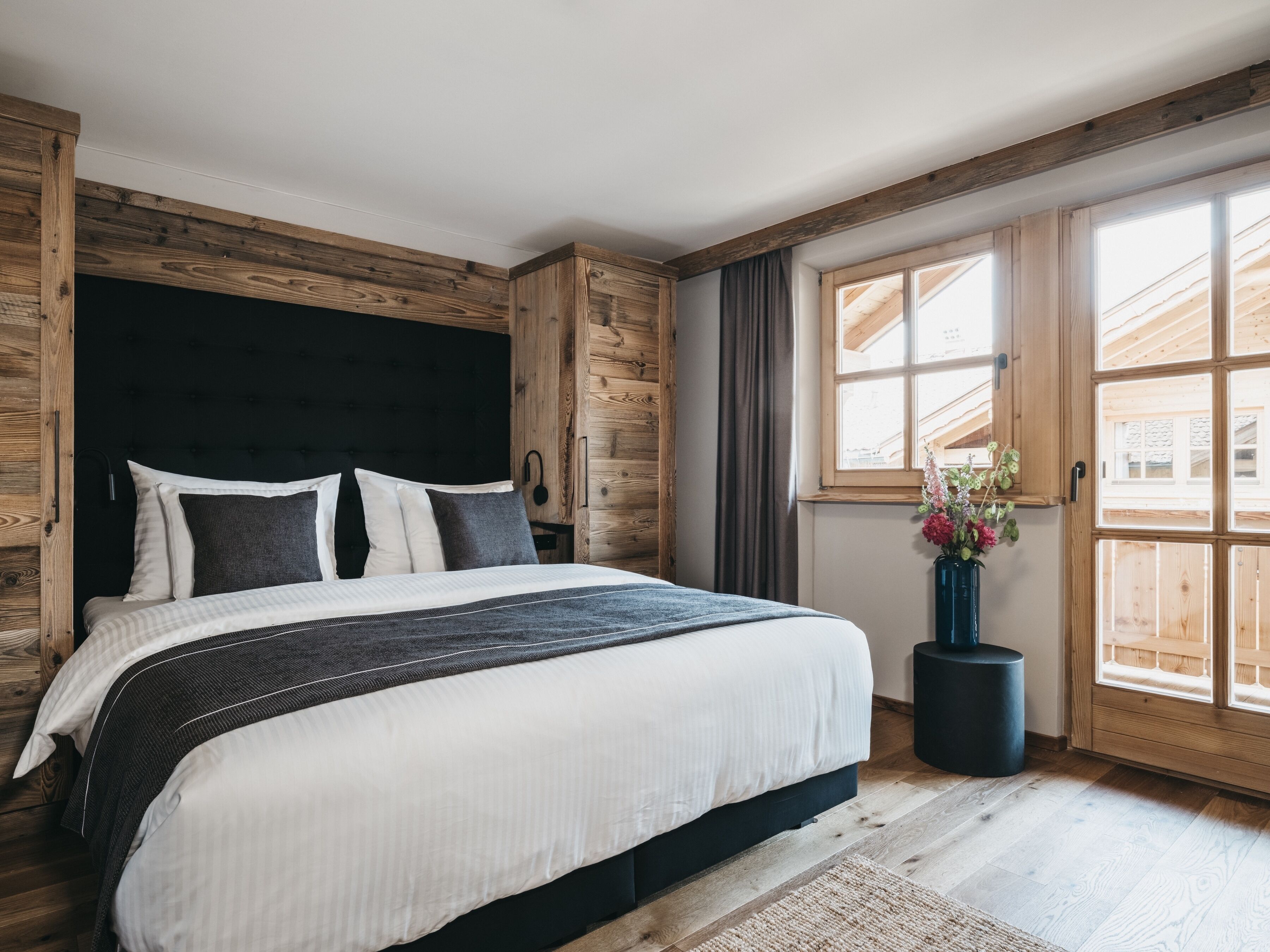 A bedroom with a large bed, wooden headboard, a window with curtains, and a vase with flowers on a side table.