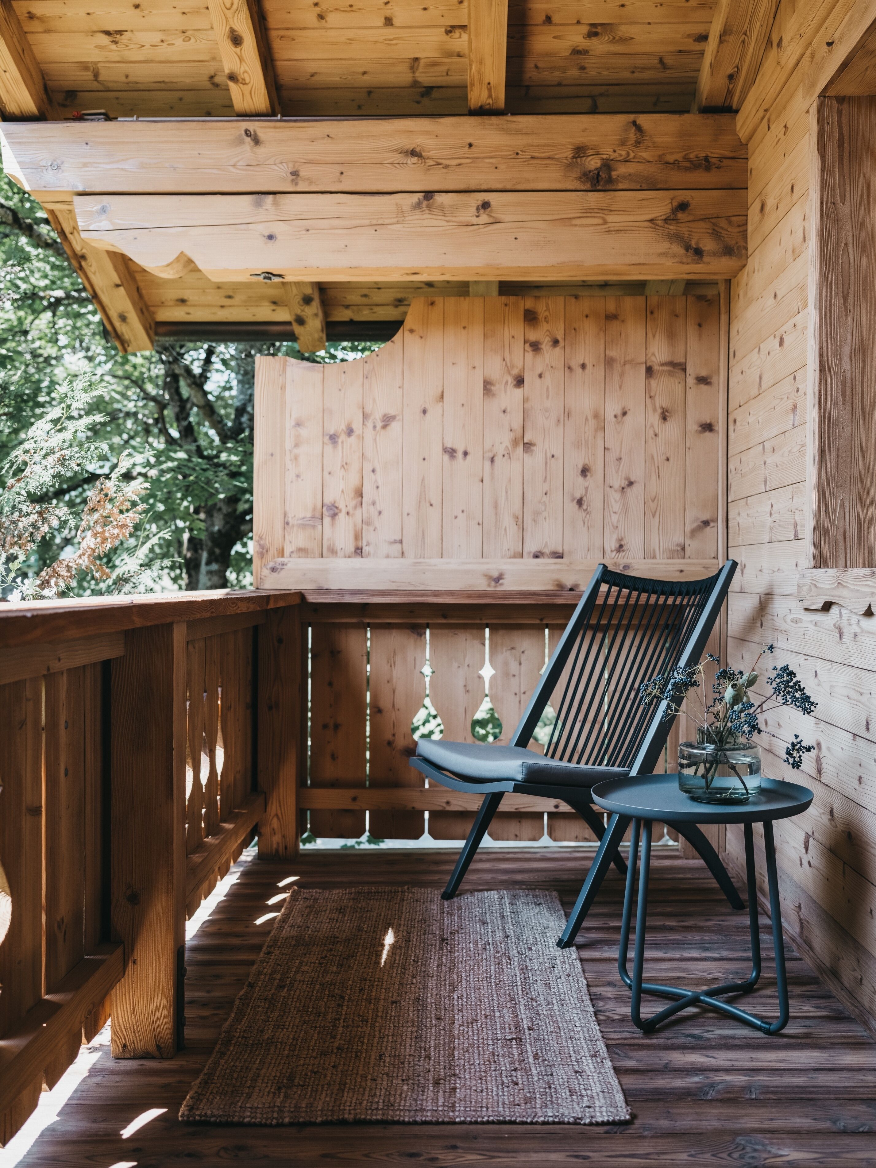 Eine Holzterrasse mit einem Stuhl, einem kleinen Tisch, einem gewebten Teppich und einem Holgitter.