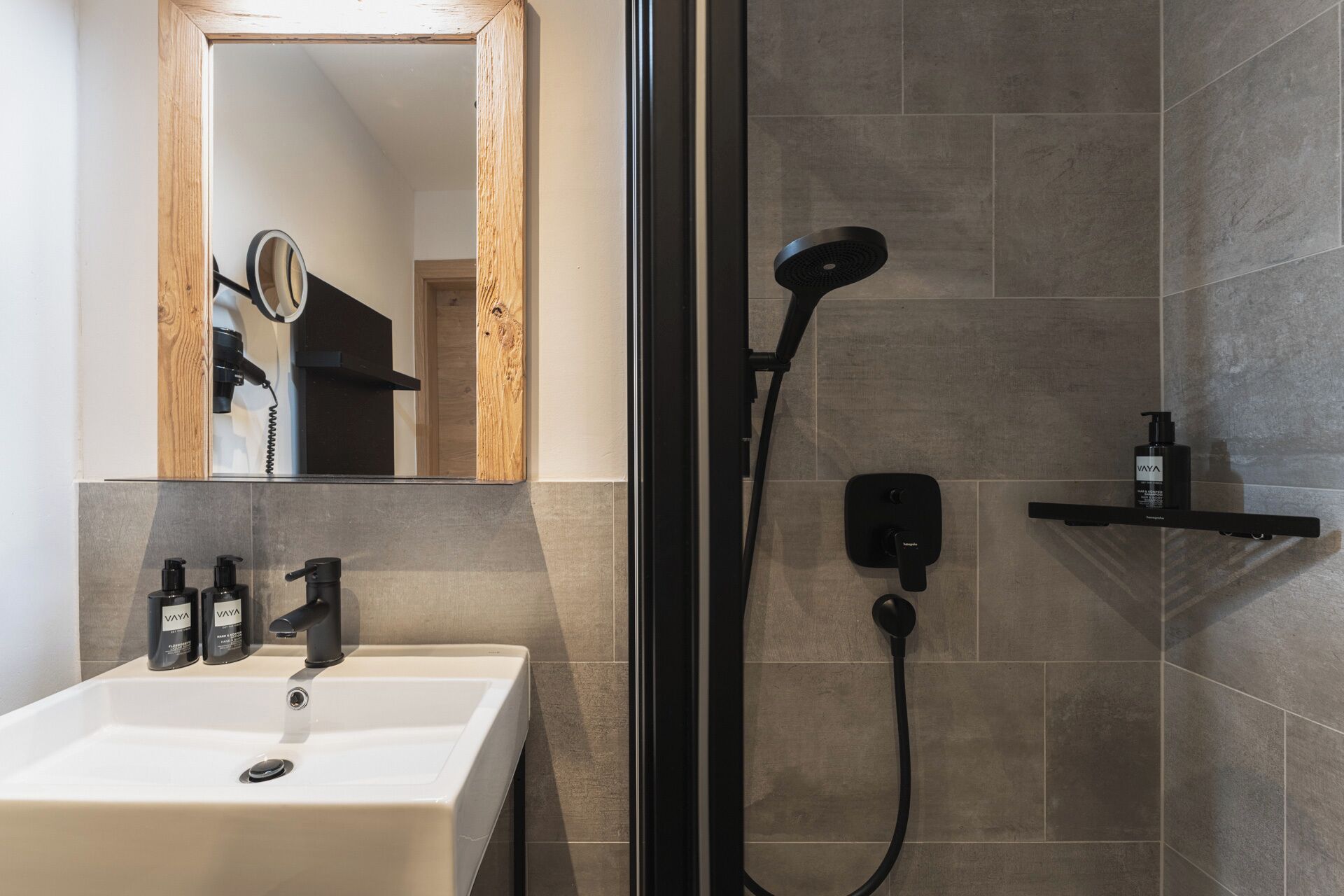 Une salle de bain avec un lavabo blanc, deux flacons de shampoing VAYA, une douchette noire à main, un téléphone noir fixé au mur et une étagère avec un flacon.