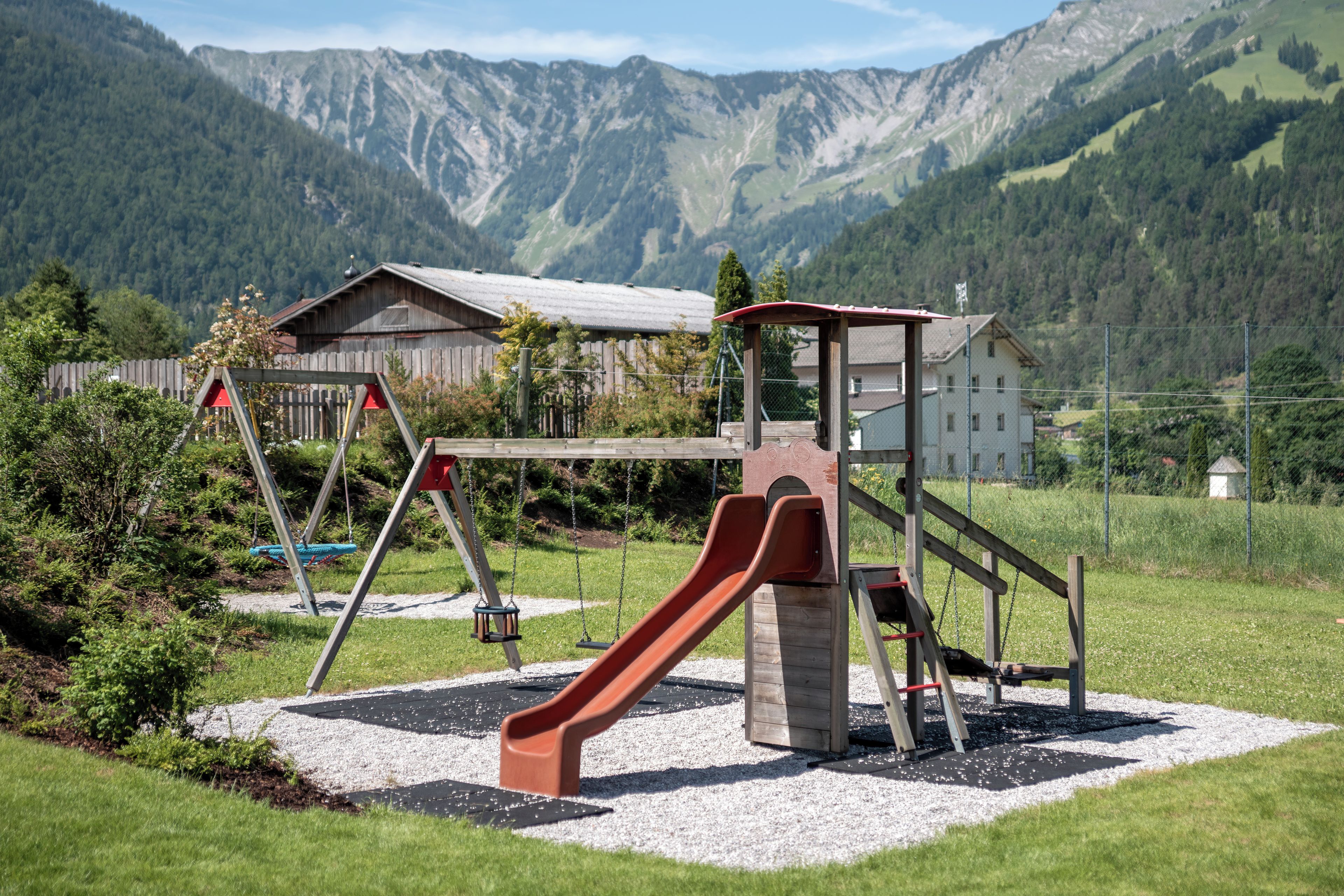 Un parque infantil con tobogán y columpios, con montañas y un edificio de fondo.