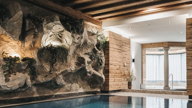 A swimming pool with a rocky wall and plants, wooden ceiling beams, and a view of the outdoors through a glass door. - VAYA Achensee (Achenkirch)