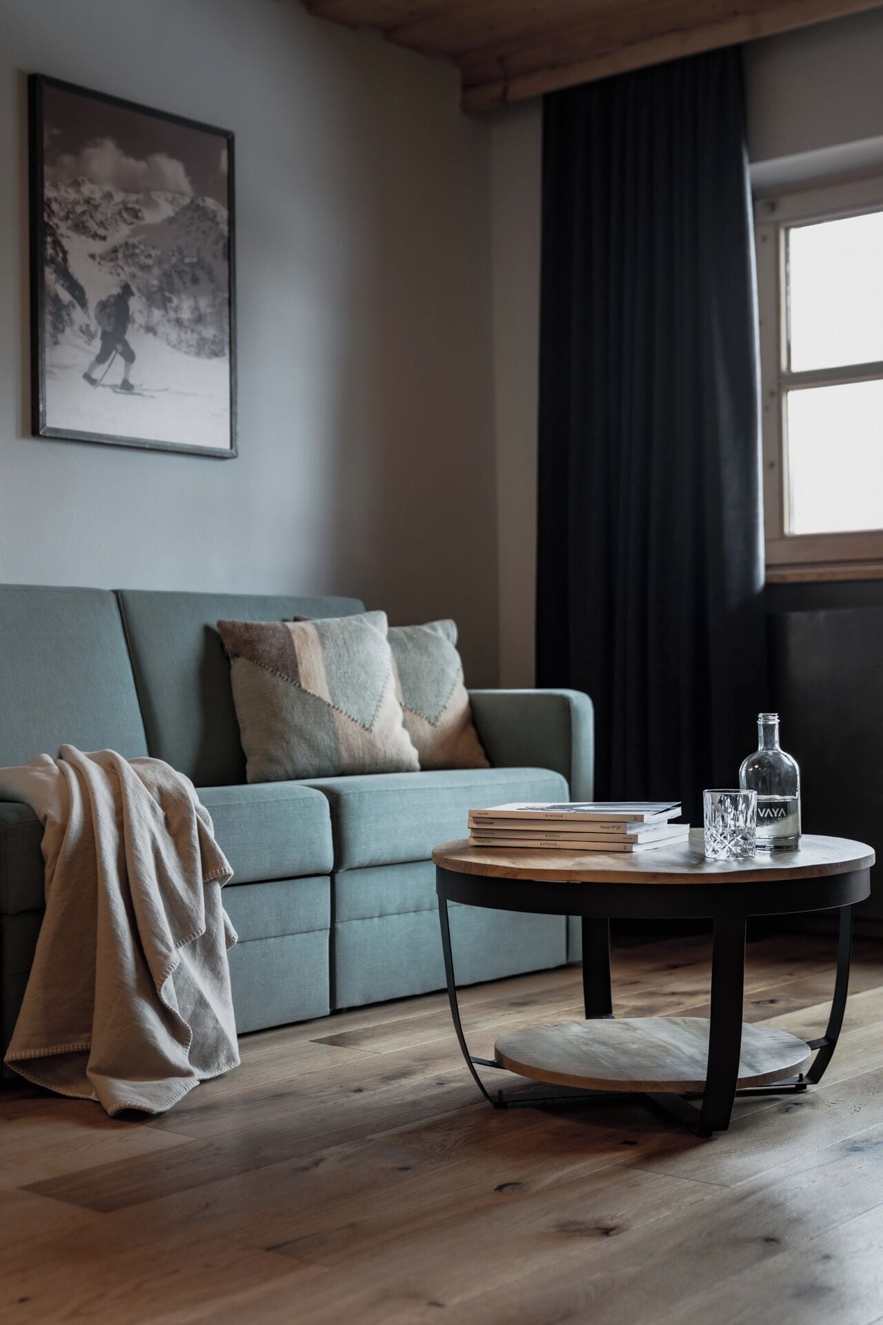 Een woonkamer met een grijze bank, een ronde salontafel met boeken en een fles, een deken op de vloer en een ingelijst schilderij aan de muur.