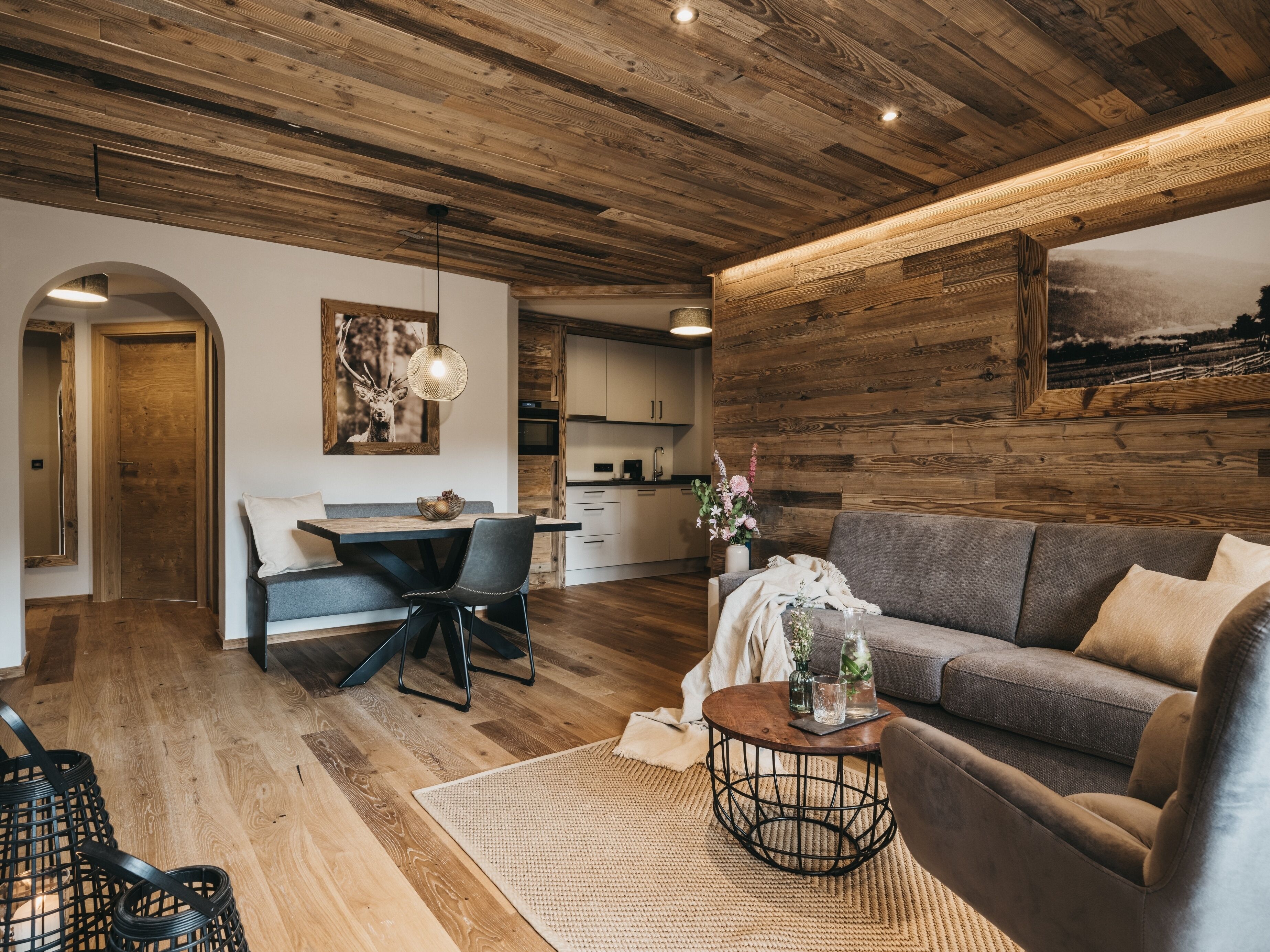Una sala de estar moderna con techo de madera, un sofá gris, una mesa de comedor con sillas y una cocina al fondo.