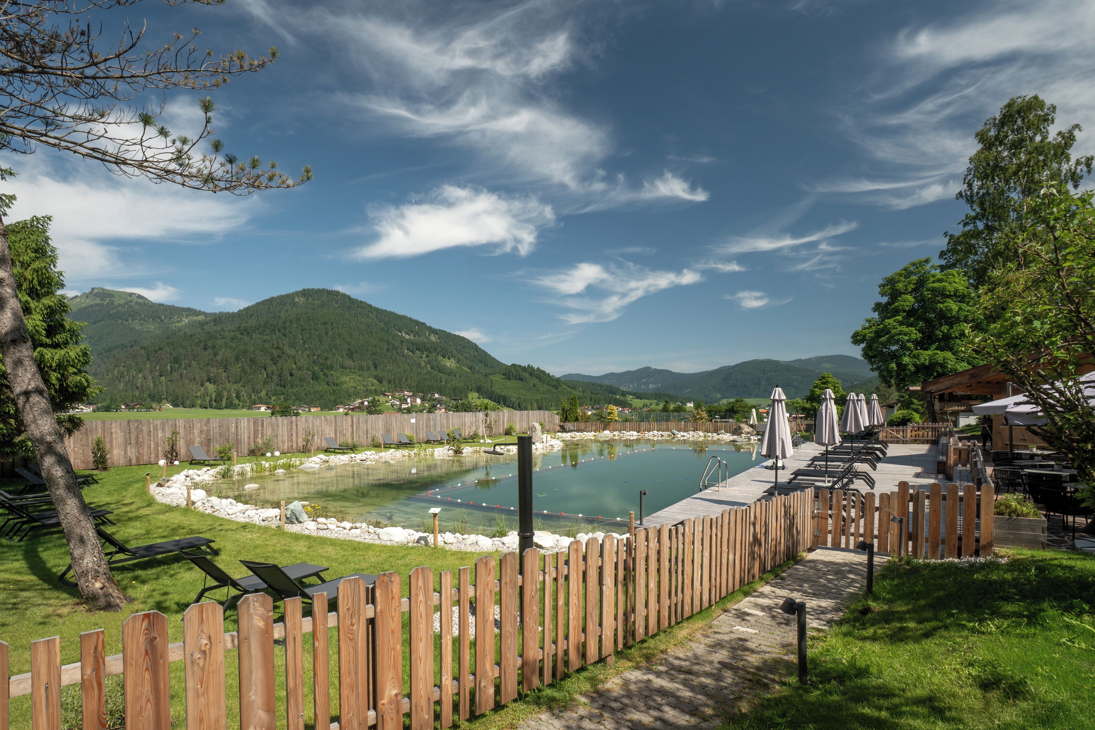 Eine Holzterrasse mit Sitzgelegenheiten im Freien, einem Schwimmbecken und Blick auf ein Gebirge.