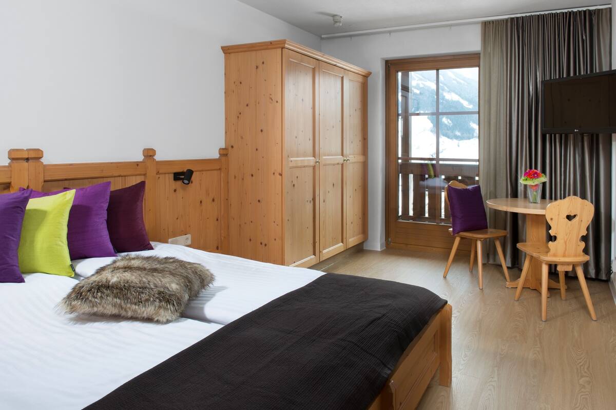 A bedroom with a bed, wooden wardrobe, a small table with a chair, and a window with a view of mountains.