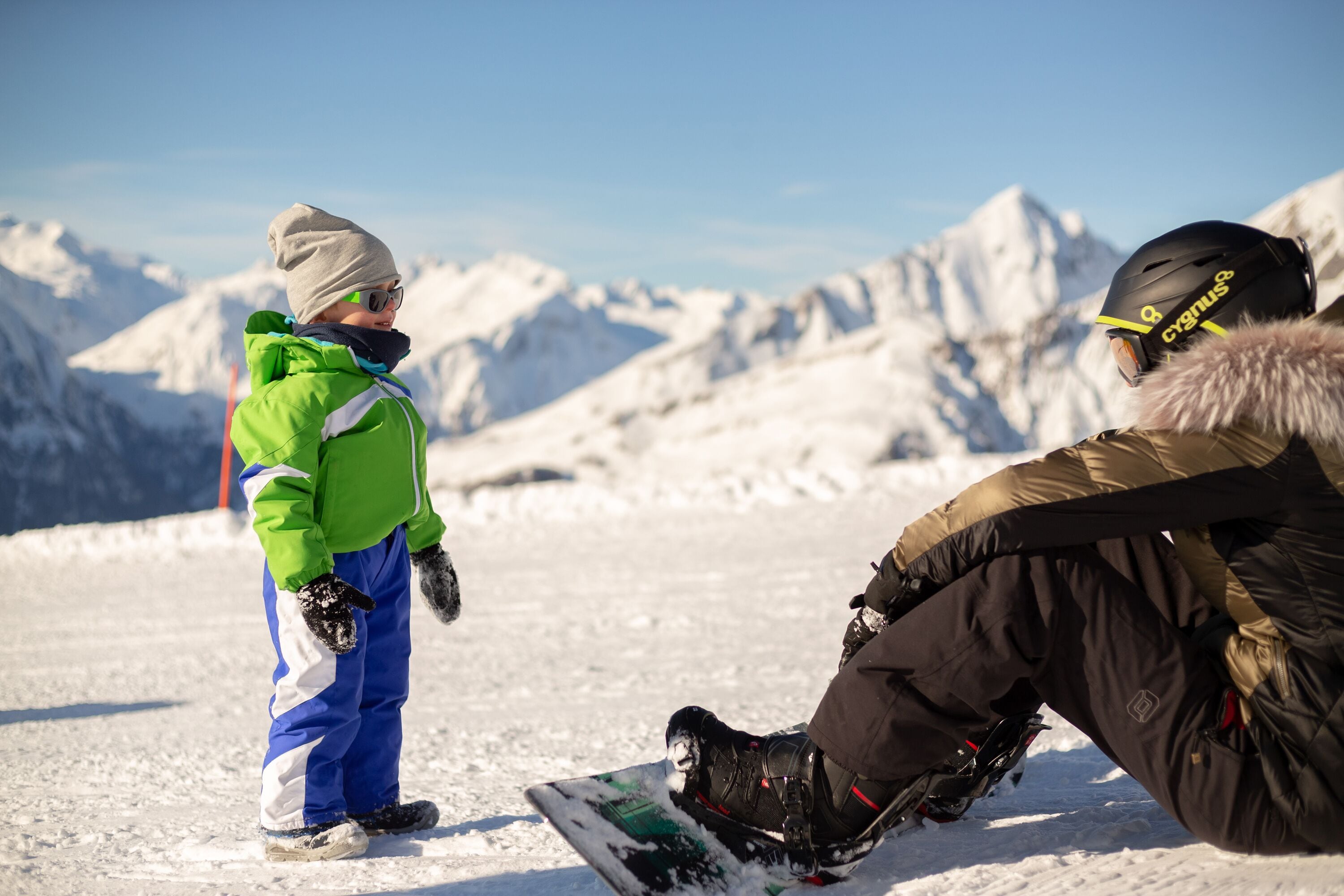 Un paisaje nevado de montañas con un snowboarder con casco negro y chaqueta marrón.