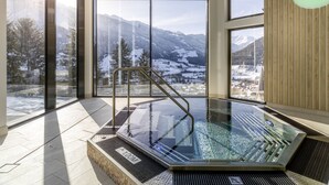 A modern indoor pool with a glass floor, large windows offering a snowy mountain view, and a metal ladder leading to the pool. - Hotel Goldried (Matrei in Osttirol)