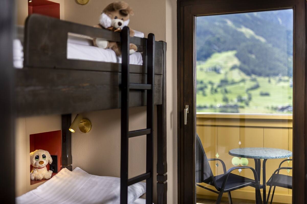 A bunk bed with a teddy bear on the top bunk and a small stuffed dog on the bottom. There is a balcony with a table and chairs, and a view of a green landscape with mountains in the distance.