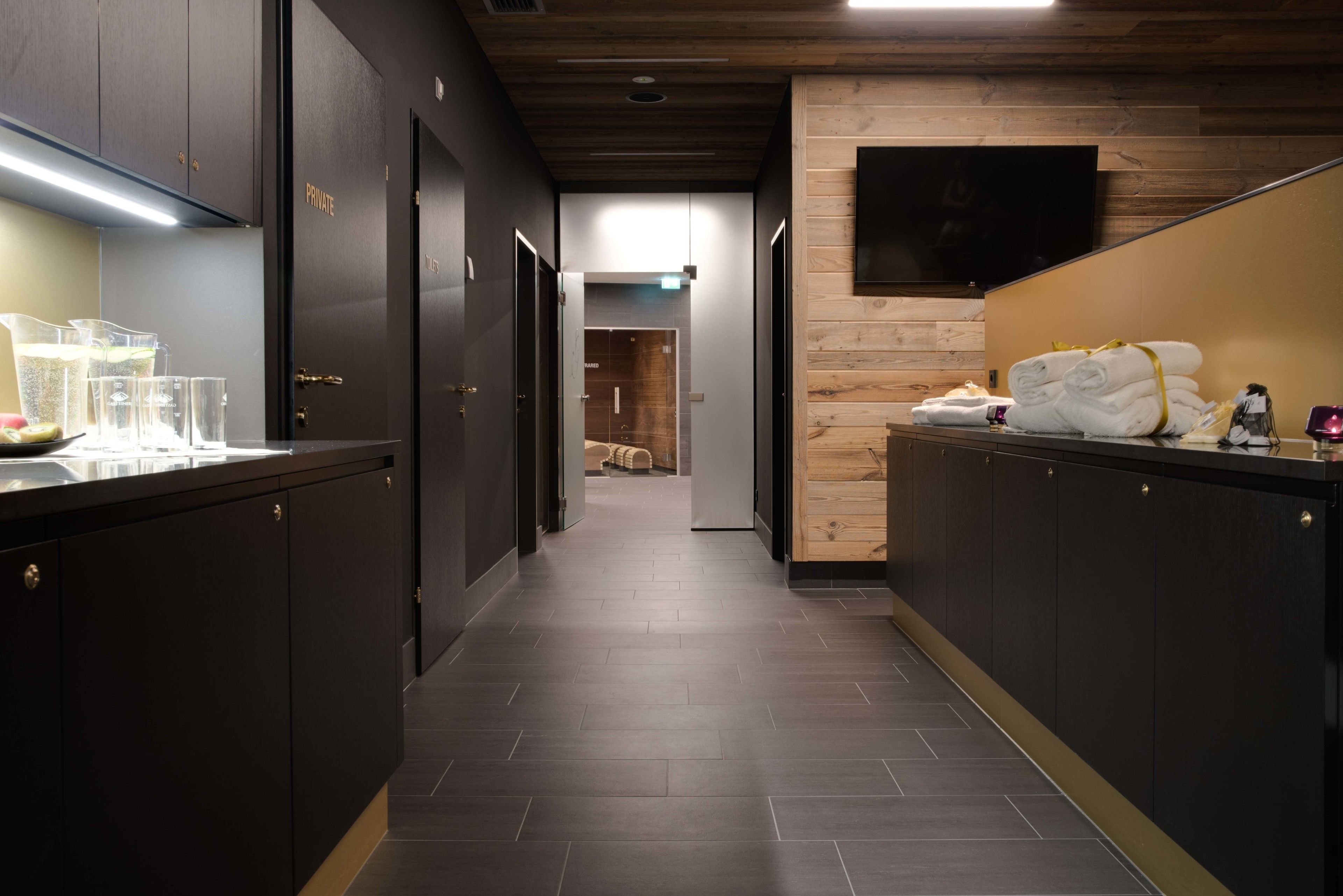 A modern hotel corridor with dark wooden cabinets, a flat-screen TV mounted on a wooden wall, and a counter with towels and toiletries.