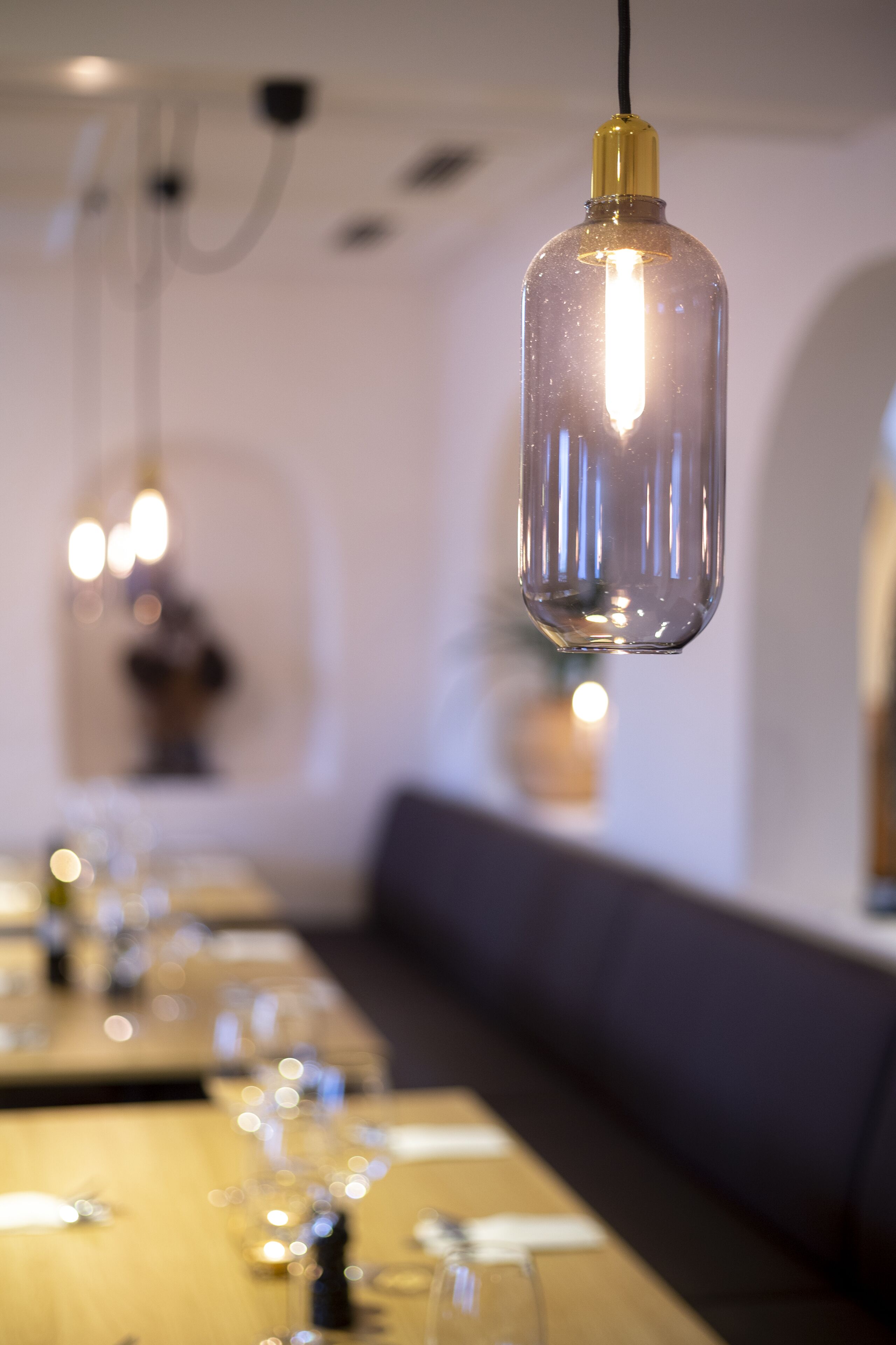 A dining area with a hanging light fixture, tables set with glasses and cutlery, and a bench along the wall.