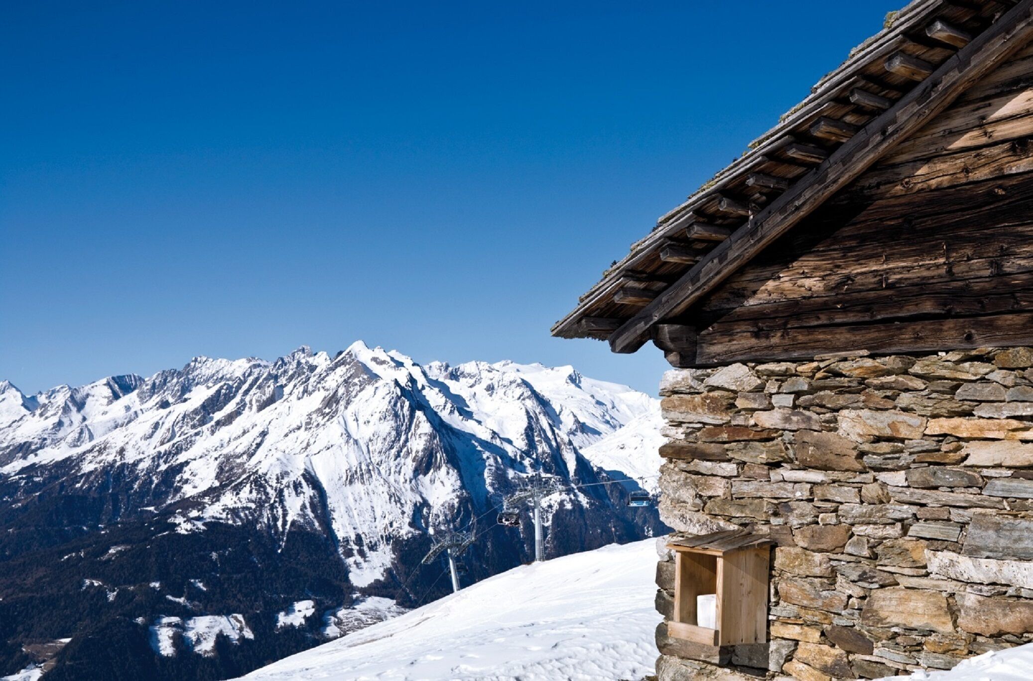 Un edificio de piedra con techo de madera, frente a un paisaje de montañas nevadas y un cielo azul despejado.